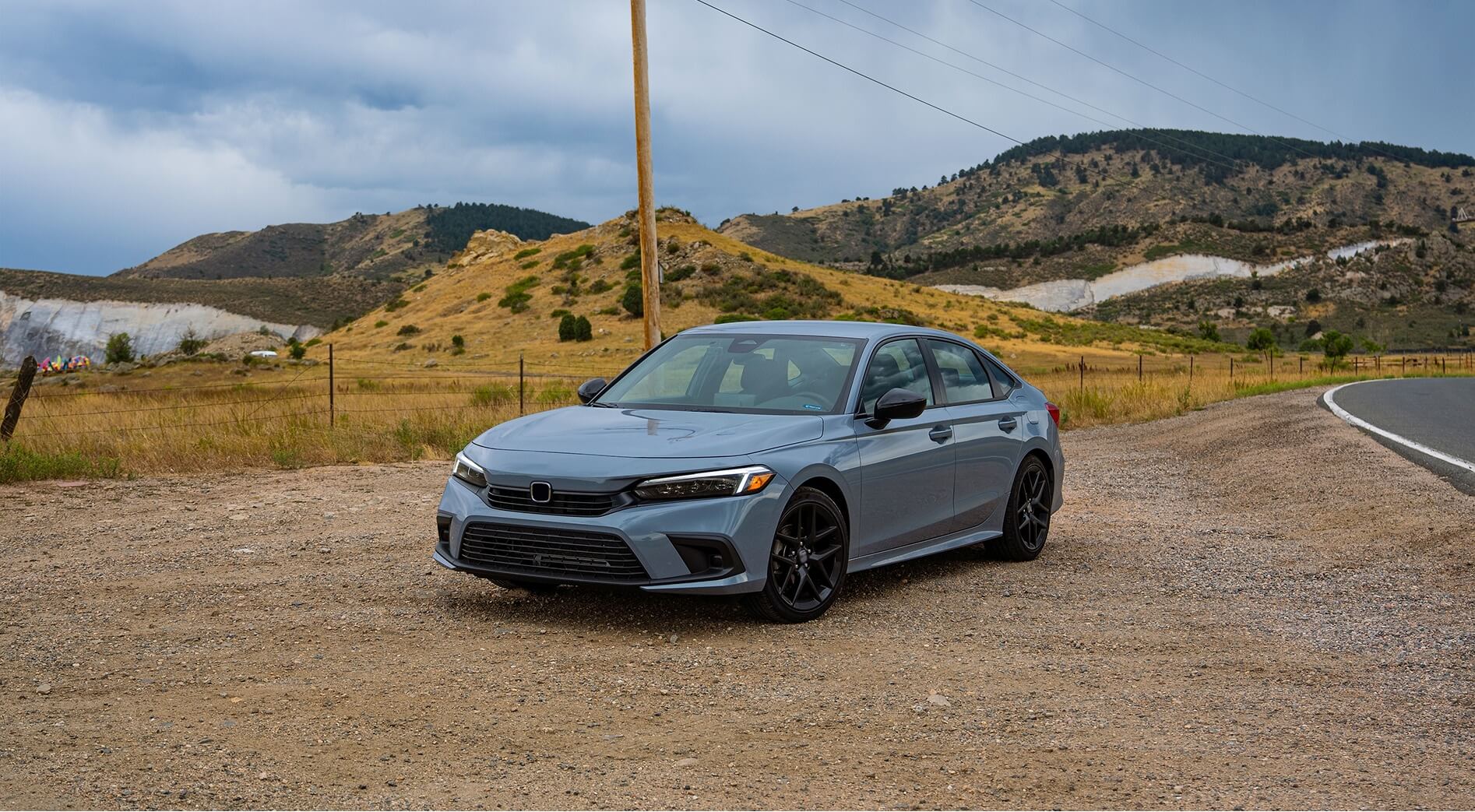 Light blue Honda Civic sedan with dark alloy wheels parked on gravel road with hills in background